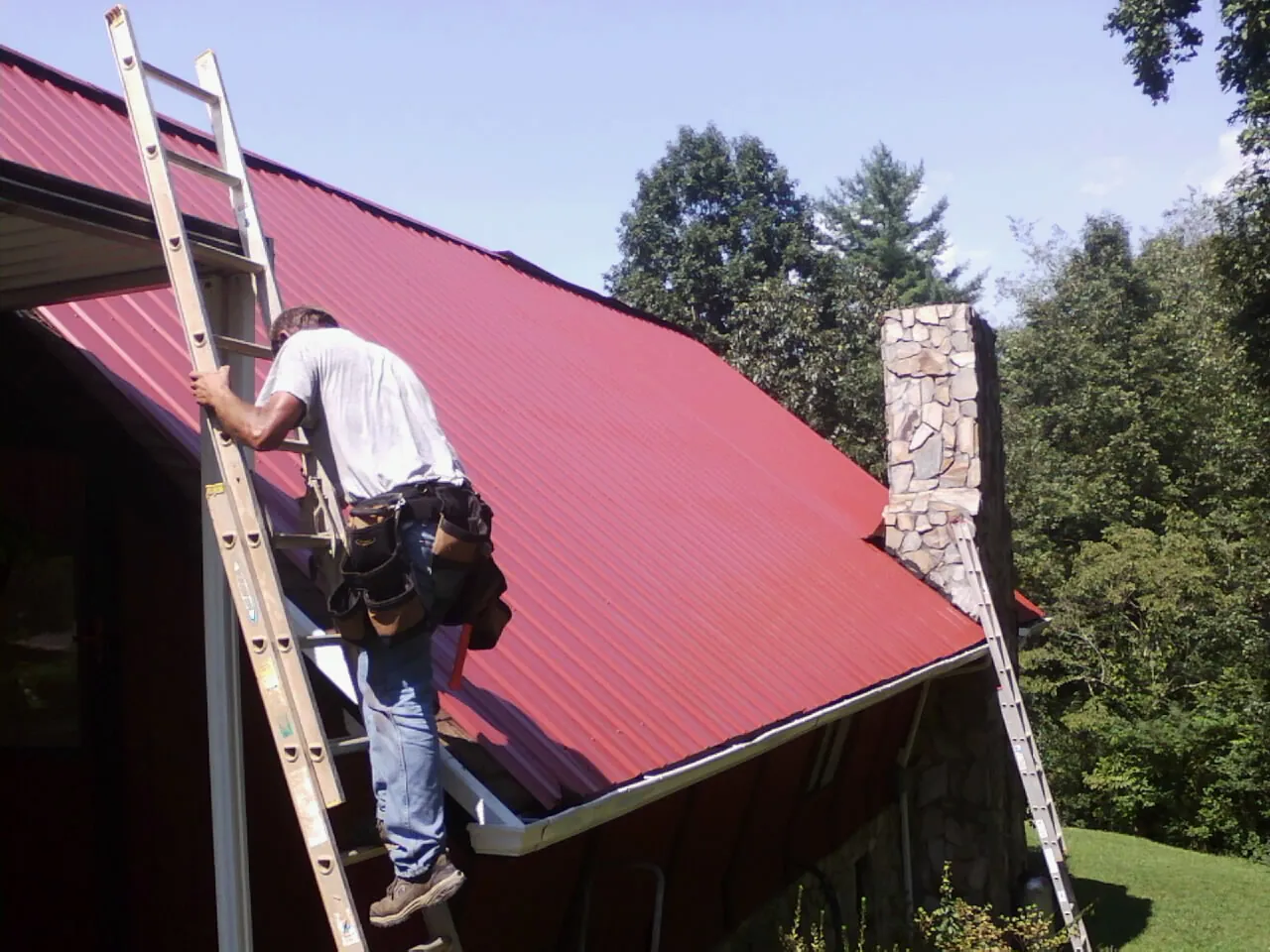 Roofer on a ladder beside a red metal roof and stone chimney — Soffit & Fascia Repair by Roofing Brothers in Vanceboro